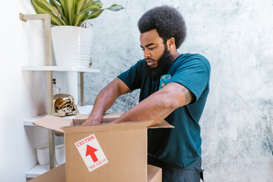 A man with dark curly hair and a beard is standing indoors, partially inside a cardboard box labeled with a red caution arrow and the text 'Caution This Side Up.' He is wearing a blue t-shirt and is actively packing or unpacking items, with his hands inside the box. Behind him, there is a white shelving unit holding decorative objects, including a large white ceramic pot with a green plant on top and a small gold-colored decorative skull. The storage area has a grey wall with a textured finish. The scene depicts logistics involved in home relocation, with the man preparing furniture or belongings for transport as part of a house removal process facilitated by Man and Van Bickley, potentially highlighting packing and loading procedures.