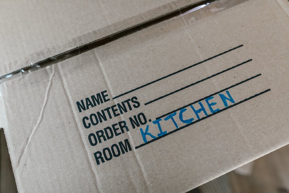 Close-up of a cardboard moving box with labels on the side, including sections for 'Name', 'Contents', 'Order No.', and 'Room', with the word 'KITCHEN' handwritten in blue ink in the 'Room' section. The box is sealed with clear packing tape and appears to be packed for a home relocation. The background shows other boxes and packaging materials, indicating an ongoing packing and moving process, likely inside or near a property doorway, with natural lighting illuminating the scene. This image reflects professional packing and logistics support offered by Man and Van Bickley for house removals, focusing on organized transport and safe handling of household items during the moving procedure.