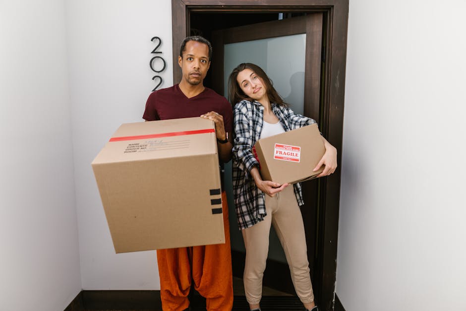 A man with dark curly hair and a beard is standing indoors, partially inside a cardboard box labeled with a red caution arrow and the text 'Caution This Side Up.' He is wearing a blue t-shirt and is actively packing or unpacking items, with his hands inside the box. Behind him, there is a white shelving unit holding decorative objects, including a large white ceramic pot with a green plant on top and a small gold-colored decorative skull. The storage area has a grey wall with a textured finish. The scene depicts logistics involved in home relocation, with the man preparing furniture or belongings for transport as part of a house removal process facilitated by Man and Van Bickley, potentially highlighting packing and loading procedures.