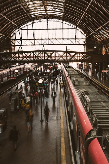 An outdoor train station platform during the late afternoon or early evening, illuminated by warm sunlight. The platform features a covered waiting area with supporting black metal columns and a roof 
