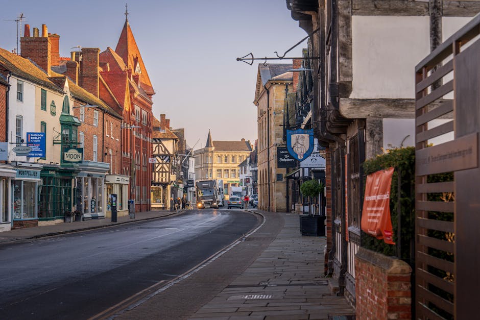 A quiet street scene in Bickley showing a variety of buildings including a brick church with a pointed spire, retail storefronts such as a shop named 'Moon' and other small businesses, and residential properties with traditional architecture. The pavement runs alongside the closed shop fronts, with a few potted plants and a wooden fence visible in the foreground on the right. Several vehicles, including a truck, are parked or moving along the curved road. The scene is captured during daylight hours with clear skies, highlighting the historic and commercial character of the area. This setting relates to home relocation and furniture transport processes that may be carried out by Man and Van Bickley as part of their house removals services.