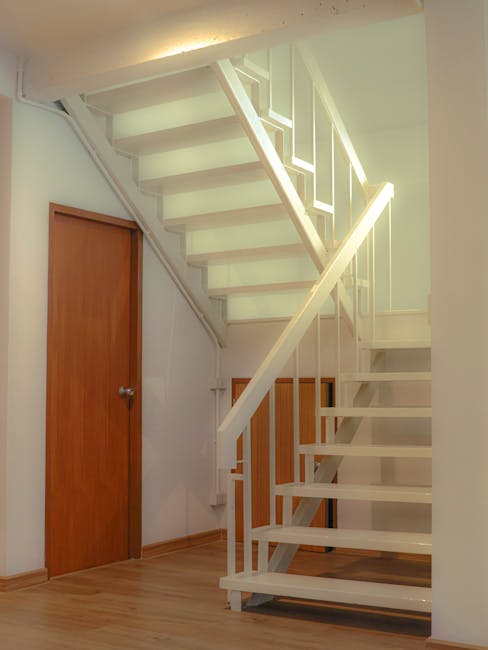 Interior view of a staircase area in a residential property, showcasing a white-painted wooden staircase with open risers and a simple rail design, leading to an upper floor. Beneath the staircase, there is a small wooden door with a round metal handle, and another larger wooden door with a similar handle is situated to the left, presumably leading to a storage closet or room. The staircase is positioned close to the wall, with a light-colored wooden floor underneath. The photo captures the tidy and functional space, likely prepared for a home relocation or furniture transport process by Man and Van Bickley, fitting the context of house removals or packing and moving services. The area is well-lit with artificial lighting, highlighting the clean and organized environment, suitable for the safe loading or unloading of boxes, packed furniture, and moving equipment.