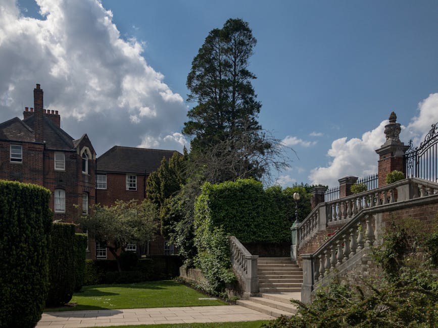 A stone staircase with decorative balustrades and ornate brick pillars leads up to a gated entrance outside a large residential property. Surrounding the staircase are well-maintained hedges, shrubs, and a tall, leafy tree, set against a backdrop of a red brick building with multiple windows and a chimney. The sky above is partly cloudy with patches of blue, and the scene is illuminated by daylight. This exterior setting depicts a typical house removal environment where furniture and boxes might be transported during a home relocation, with the staircase and garden area serving as part of the moving process handled by professionals like Man and Van Bickley.