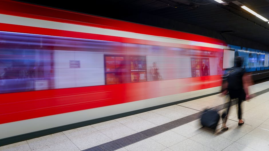 A blurred underground train with a red and white exterior is arriving or departing at Bickley Station, with a person dressed in dark clothing walking along the platform pulling a wheeled suitcase. The platform surface is light-colored tile, and overhead lighting illuminates the scene. Inside the station, reflections and faint outlines of shelves and signs are visible through the train's windows, indicating it is a busy commuter environment. This image captures the dynamic movement typical of home relocation or packing and moving activities, with [COMPANY_NAME] providing essential logistical support for such furniture transport and train link moves associated with Bickley Station, as referenced in the guide for easy train link moves.