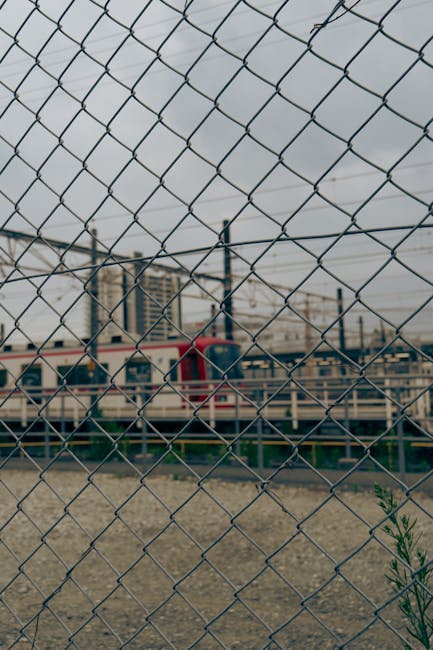 A view through a metal chain-link fence with diamond-shaped mesh pattern, showing a train parked on railway tracks in an urban environment. The train has a red and white colour scheme and is partially obscured by the fence. In the background, there are modern multi-storey buildings, possibly apartments or offices, under an overcast sky with grey clouds. The ground inside the fenced area appears to be gravel or dirt, with some sparse greenery visible at the bottom right corner. This scene suggests an urban setting where local house removals or furniture transport might involve navigating railway lines or train stations, aligning with the services offered by Man and Van Bickley, especially around Bickley station for smooth train link moves.