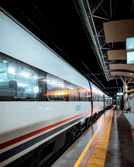 A blurred underground train with a red and white exterior is arriving or departing at Bickley Station, with a person dressed in dark clothing walking along the platform pulling a wheeled suitcase. The platform surface is light-colored tile, and overhead lighting illuminates the scene. Inside the station, reflections and faint outlines of shelves and signs are visible through the train's windows, indicating it is a busy commuter environment. This image captures the dynamic movement typical of home relocation or packing and moving activities, with [COMPANY_NAME] providing essential logistical support for such furniture transport and train link moves associated with Bickley Station, as referenced in the guide for easy train link moves.