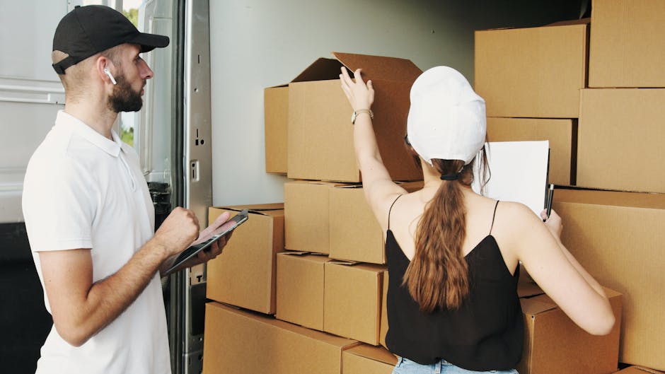 A man and a woman are engaged in a home relocation process involving packing and furniture transport. The man, wearing a white t-shirt, black cap, and wireless earbuds, is standing to the left, holding a clipboard and pen, likely taking notes or checking the packing list. The woman, with long brown hair tied back and wearing a white protective cap and black spaghetti strap top, is on the right, placing a cardboard box into a van through an open side door. The scene takes place next to a driveway or loading area with several stacked cardboard boxes and packing materials visible inside the van. The lighting is natural, highlighting the organized loading process essential for safe and efficient moving logistics. This image exemplifies proper handling and packing during a home removal, as handled by a professional service such as Man and Van Bickley, which specialises in removals and furniture transport. Capturing the careful planning and execution of a household move, it underscores the importance of proper packing materials and systematic loading, aligning with best practices in moving services.
