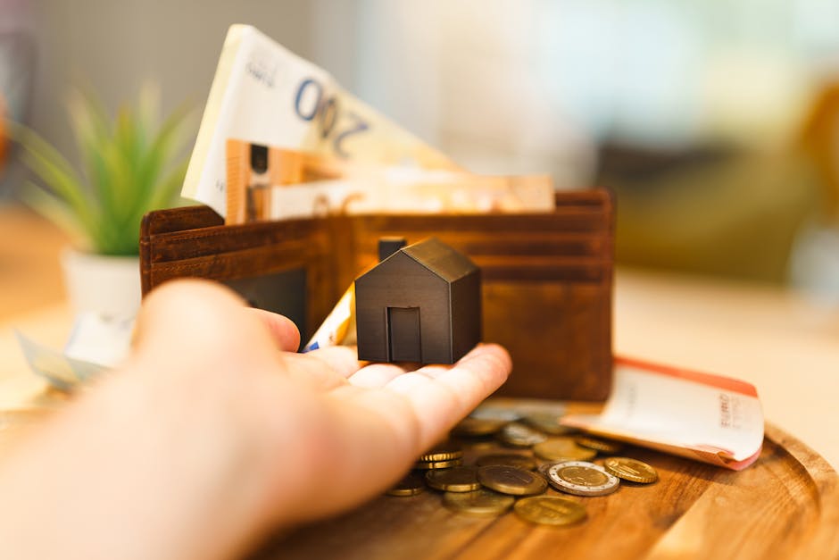A close-up view of a person's hand holding a small black house-shaped keyring, positioned in front of a brown leather wallet that is partially open, revealing several neatly stacked 20-pound and 50-pound banknotes inside. The wallet is resting on a wooden surface, with loose coins scattered around, including some silver and gold-colored currency. In the background, there is a blurred potted plant with green foliage, suggesting a domestic setting. This image captures elements of home financial management and planning related to house removals and moving logistics, as presented on the page 'BR1 removals pricing explained for Bickley homeowners' at manandvanbickley.co.uk, representing the importance of budget considerations during a house relocation with services by Man and Van Bickley.