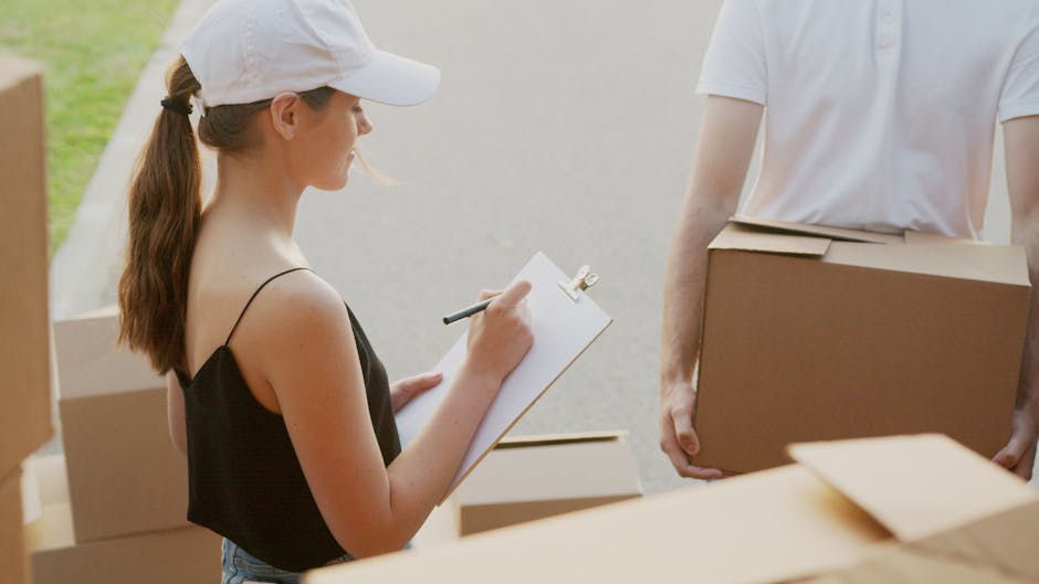A young woman with long brown hair tied in a ponytail, wearing a white cap and a black sleeveless top, is standing outdoors during a home relocation process. She is holding a clipboard with a pen and