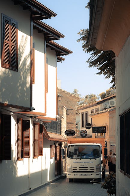 A small narrow street in Bickley with traditional two-storey houses featuring white exterior walls, brown wooden shutters on the windows, and tiled roofs. A white moving van from Man and Van Bickley is parked on the pavement, partially blocking the view of the street. The van is positioned perpendicular to the houses, with the front facing toward the camera, indicating an active furniture transport and home relocation process. A person is visible near the van, possibly involved in loading or unloading items such as cardboard boxes, wrapped furniture, or packing materials. The scene is set during daylight with clear skies, and small trees and shrubs are visible above the buildings, emphasizing the residential setting. This outdoor environment illustrates the logistics involved in professional removals for Bickley homes and gardens, with the van ready to facilitate a packing and moving operation as part of a comprehensive house removals service provided by Man and Van Bickley.