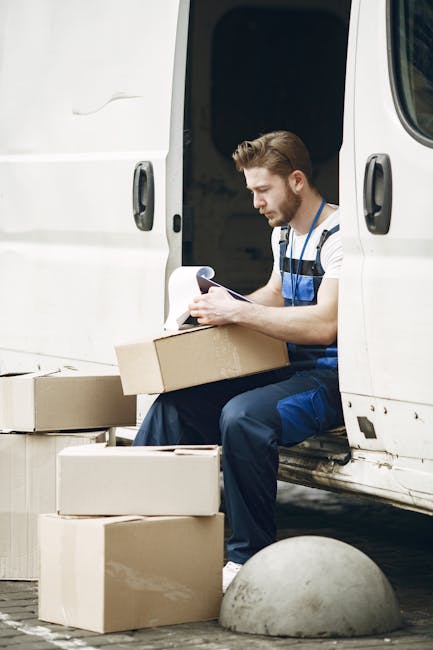 A man with short brown hair and a beard, dressed in a striped T-shirt and blue work overalls, is seated at the back of a white moving van with the sliding door open. He is holding and reading a document, likely a packing list or delivery instruction. In front of him on the ground are several cardboard boxes of various sizes, some stacked and others placed on the pavement. The boxes appear to be sealed with packing tape, and some are open, revealing packing materials inside. The van is situated on a paved area near a curb, with a rounded concrete bollard visible in the foreground. The environment is well-lit, suggesting daytime, and the scene depicts the process of home relocation, furniture transport, or packing and moving activities, as part of a house removal service. This image exemplifies items being prepared for loading or unloading during a house move facilitated by Man and Van Bickley, known for same-day removals in Bickley.
