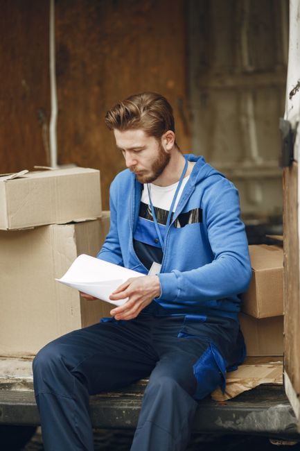 A man with short brown hair and a beard, dressed in a striped T-shirt and blue work overalls, is seated at the back of a white moving van with the sliding door open. He is holding and reading a document, likely a packing list or delivery instruction. In front of him on the ground are several cardboard boxes of various sizes, some stacked and others placed on the pavement. The boxes appear to be sealed with packing tape, and some are open, revealing packing materials inside. The van is situated on a paved area near a curb, with a rounded concrete bollard visible in the foreground. The environment is well-lit, suggesting daytime, and the scene depicts the process of home relocation, furniture transport, or packing and moving activities, as part of a house removal service. This image exemplifies items being prepared for loading or unloading during a house move facilitated by Man and Van Bickley, known for same-day removals in Bickley.